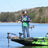 An angler in a colorful fishing jersey stands on the deck of a bass boat, holding a fishing rod