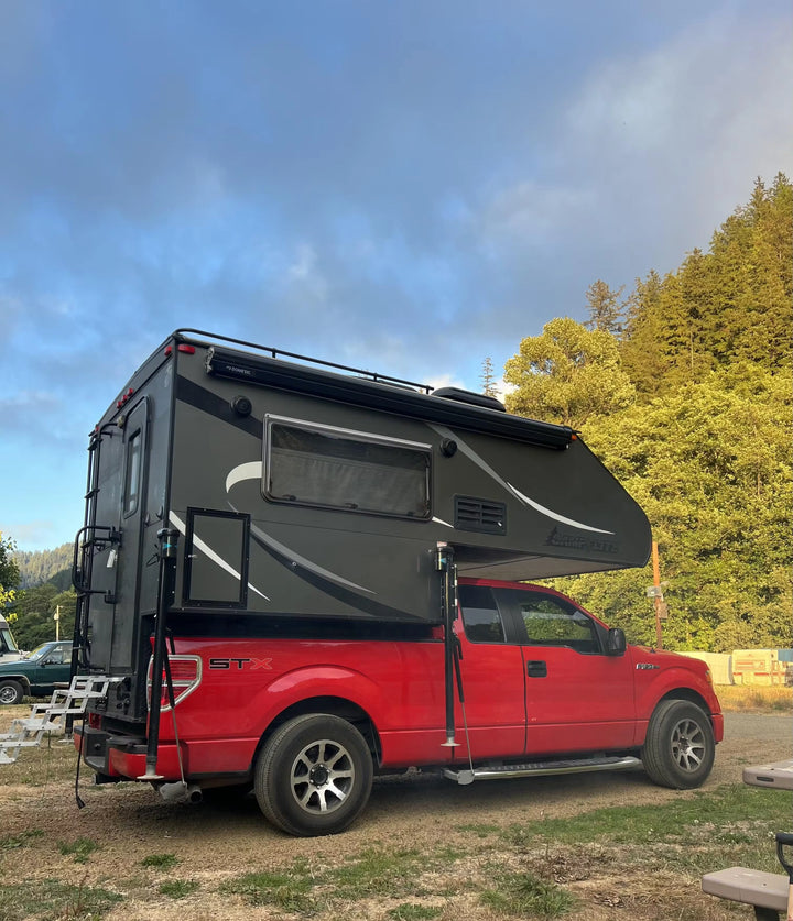 Red pickup truck with a large black camper pod attached, parked in a grassy field near woods